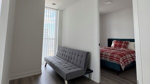 A modern bedroom with a grey sofa, a bed with a red and white checkered bedspread, and a large window with blinds. - Cozy Condo in Vaughan (Vaughan)