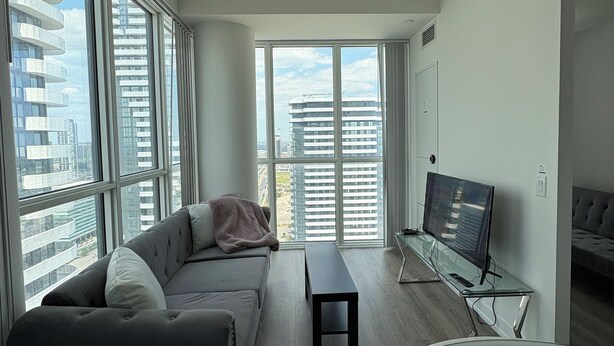 A modern living room with a sofa, a glass coffee table, a flat-screen TV, and large windows offering a city view. - Cozy Condo in Vaughan (Vaughan)