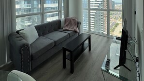 A modern living room with a grey sofa, a glass coffee table, and a TV on a stand. Large windows offer a view of high-rise buildings. - Cozy Condo in Vaughan (Vaughan)