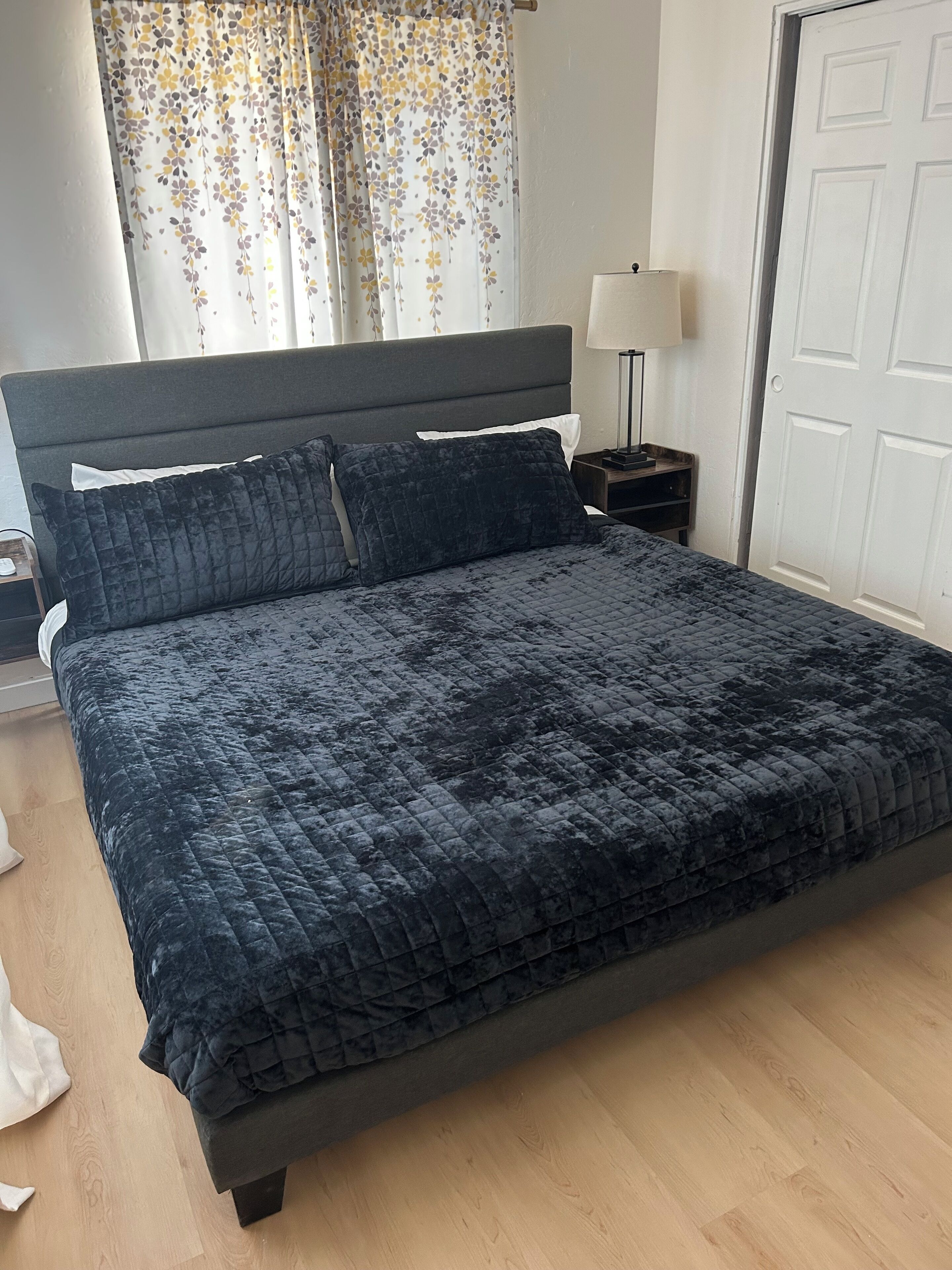 A bed with a dark quilted cover, a bedside table with a lamp, and a door in the background.