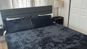 Una cama con una colcha acolchada oscura, una mesita de noche con una lámpara y una puerta al fondo.