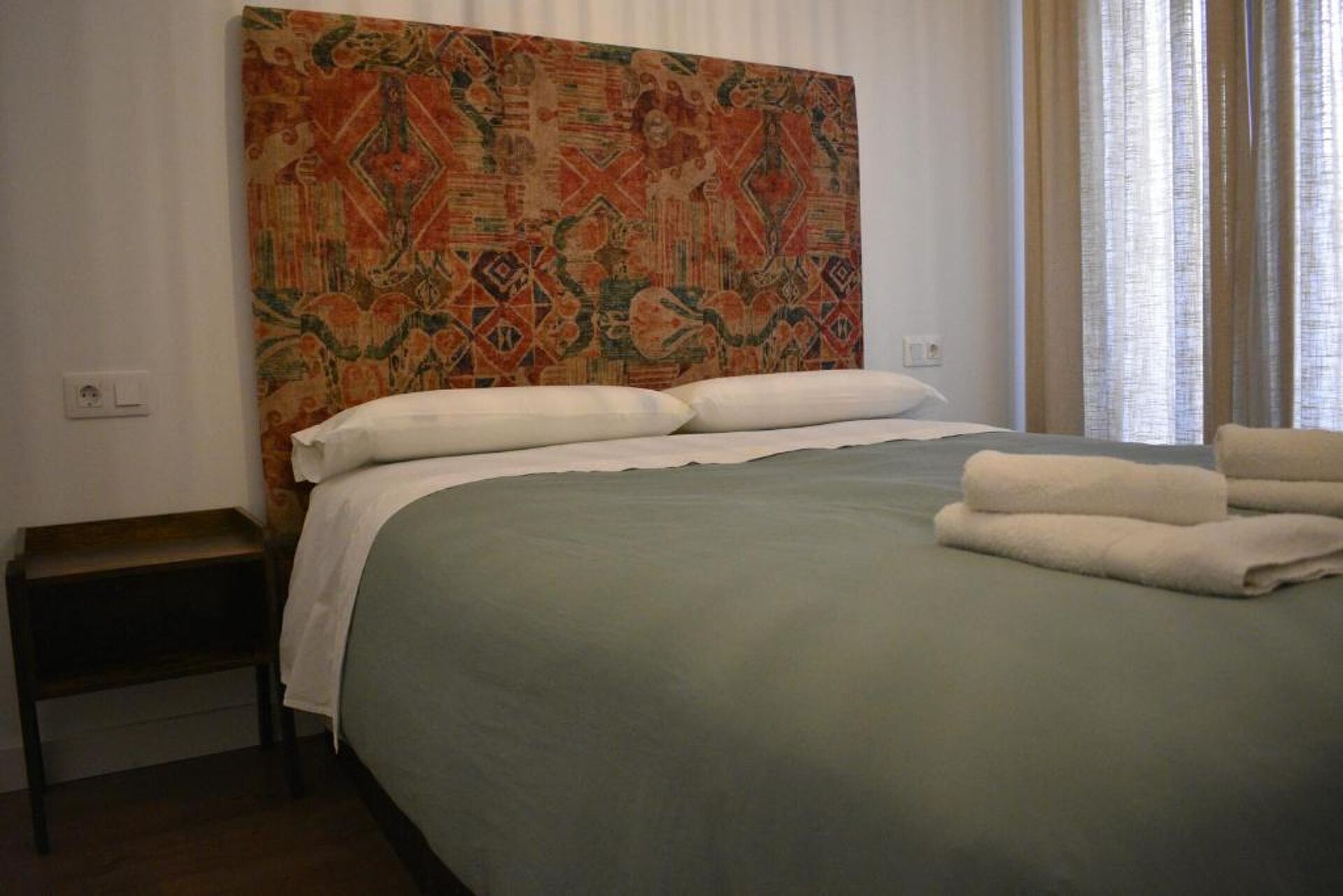 Un lit bien fait, avec une couvre-lit verte et des oreillers blancs, une table de chevet en bois recouverte d’un tissu blanc, et une tapisserie colorée accrochée à la tête de lit.