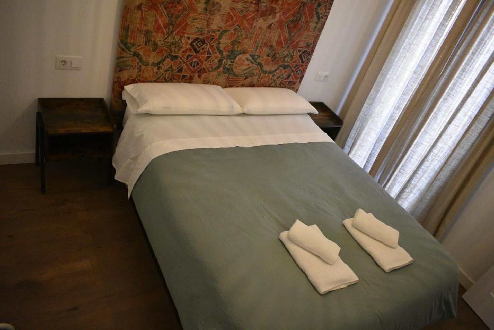 Una cama bien hecha con una colcha verde y dos almohadas, una mesita de noche de madera y un tapiz en la pared.