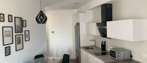 Une cuisine moderne avec des meubles blancs, un four à micro-ondes et un évier. On y trouve également un coin repas avec une table en verre et des chaises vertes.