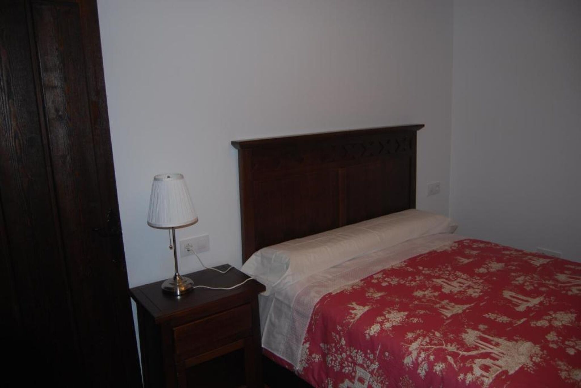 A bedroom with a wooden headboard, a bedside table with a lamp, and a red patterned bedspread.