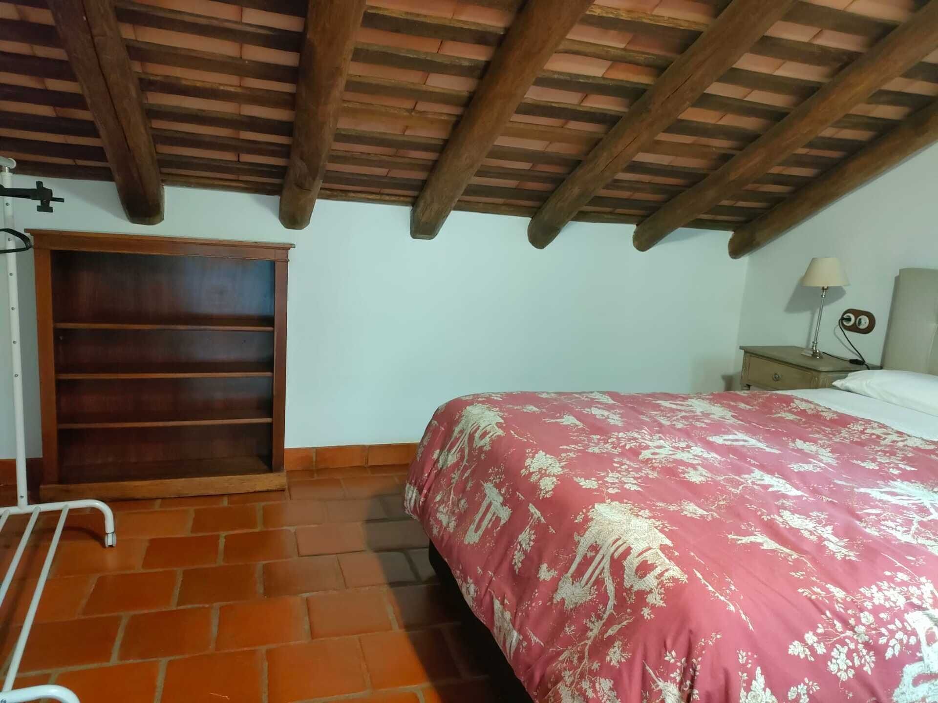 A bedroom with a wooden ceiling, a bed with a floral bedspread, a nightstand with a lamp, and a wardrobe.