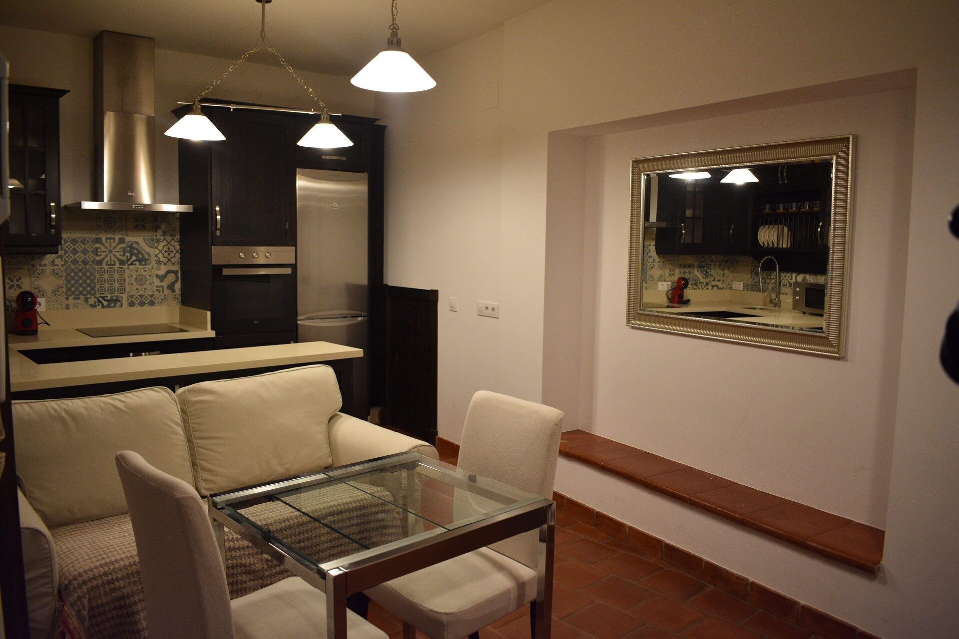 Una cocina moderna con armarios oscuros, una campana extractora de acero inoxidable y un horno empotrado. Hay una zona de comedor con una mesa de vidrio y sillas blancas.