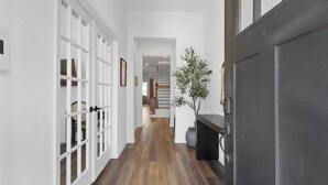 A hallway with white walls, wooden flooring, and a dark door. There is a potted plant and a framed picture on the wall. - Houston Family Retreat | Sleeps 10 | Near NRG (Houston)