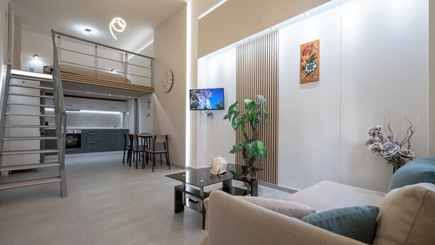 A modern living room with a sofa, a coffee table, and a TV. There is a kitchen area with a wooden ceiling and a staircase.