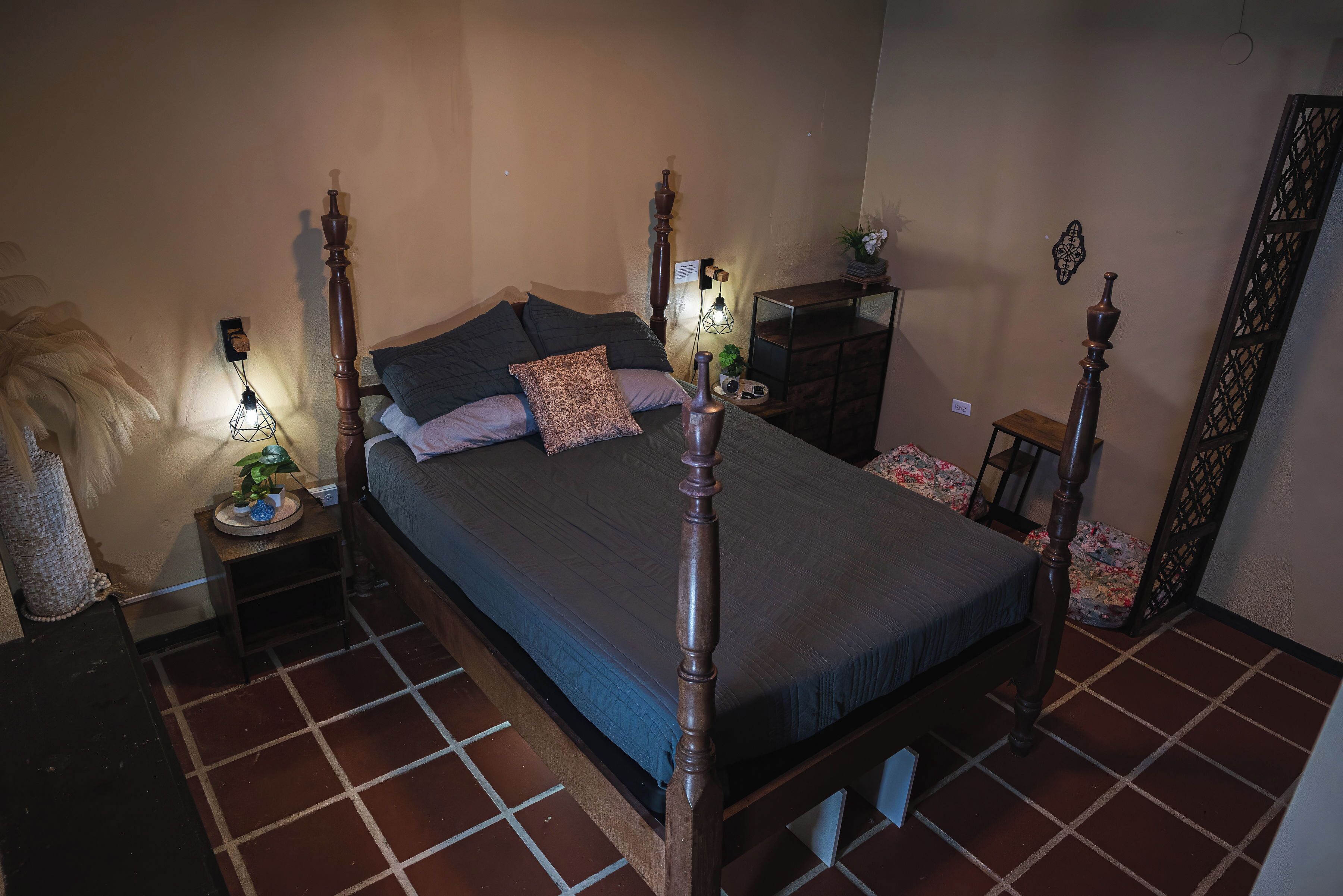 A bedroom with a four-poster bed, a nightstand with a lamp, a dresser, and a mirror.