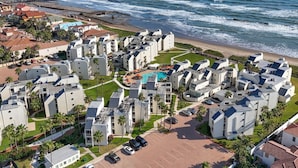 A coastal residential area with modern buildings, a central swimming pool, and a pier in the background.