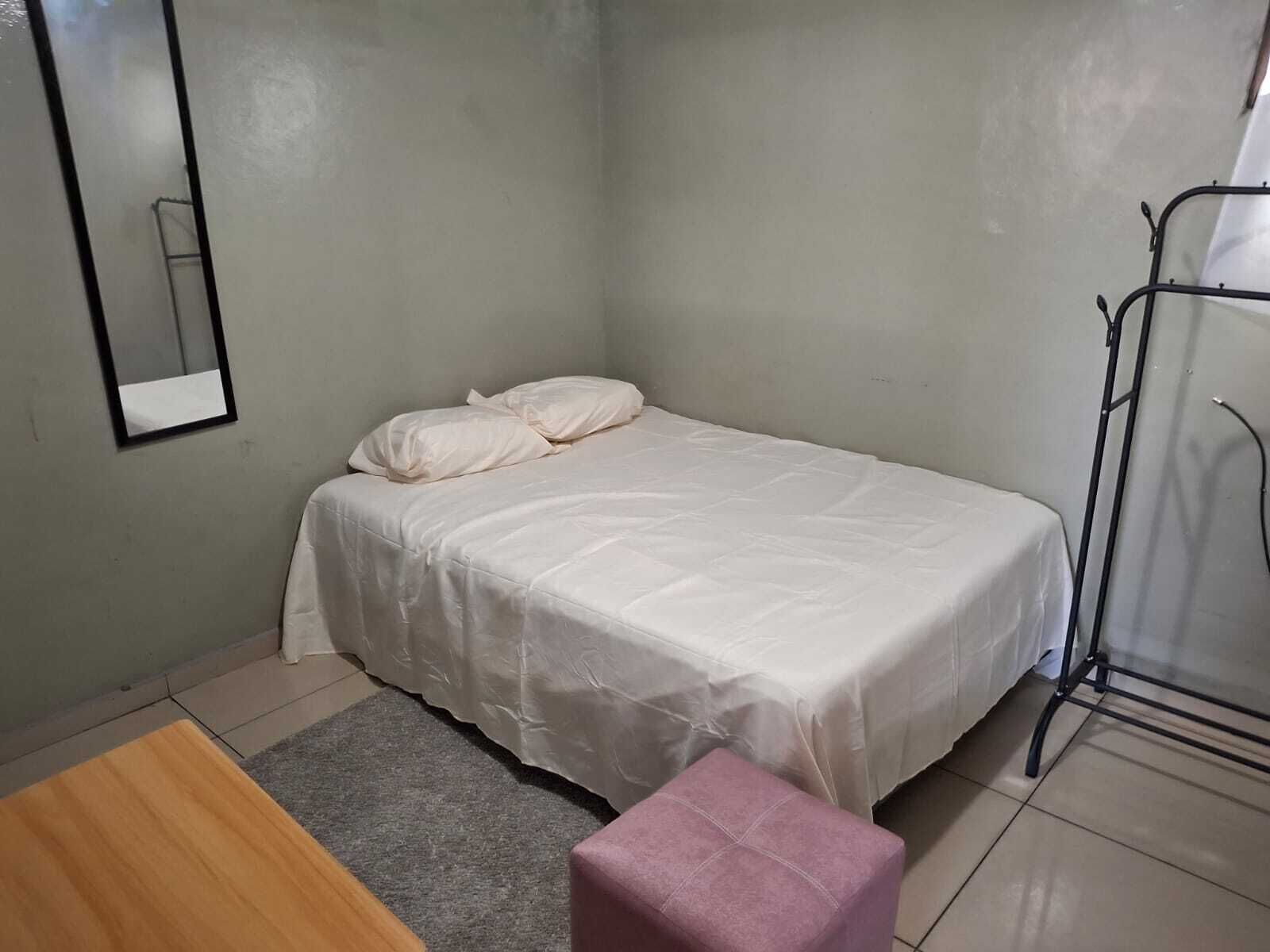 Ein kleines, ordentliches Schlafzimmer mit einem Einzelbett, einem Holztisch, einem rosafarbenen Hocker und einem Wäscheständer.