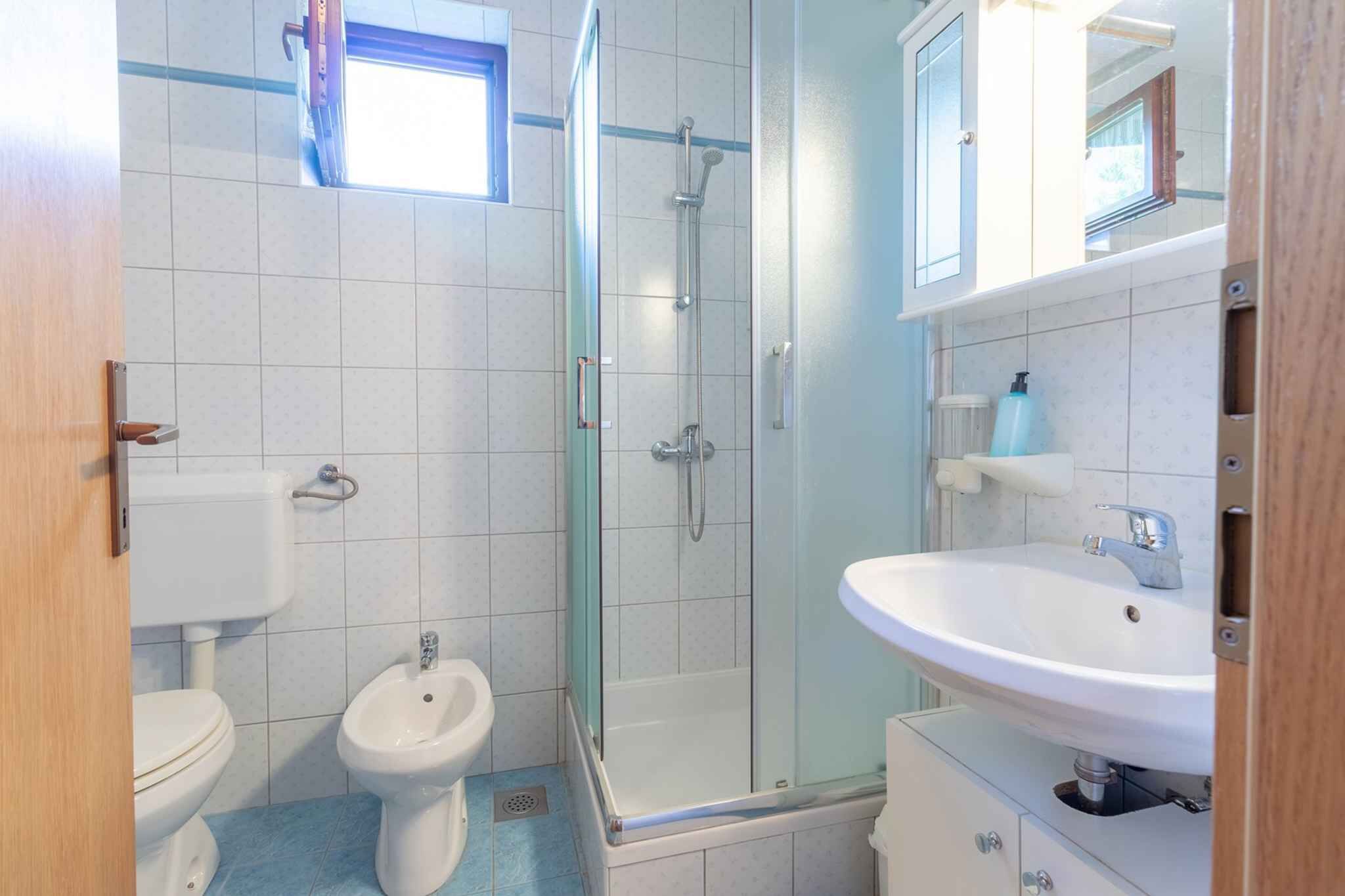 A bathroom with a toilet, bidet, glass shower enclosure, sink, and a window above the toilet.
