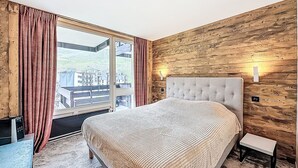 A bedroom with a large bed, wooden walls, a view through a window, and a small bedside table.