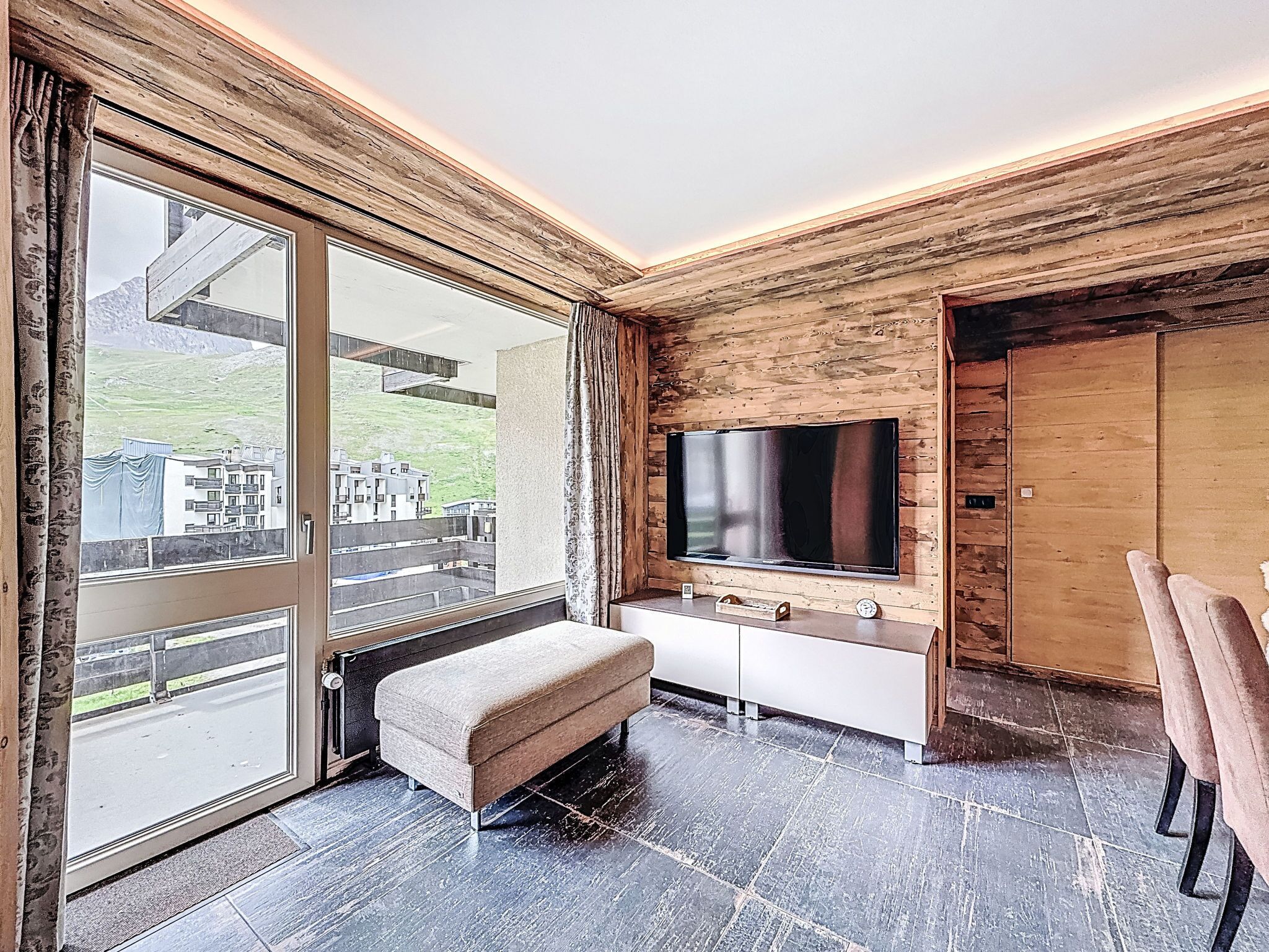 A modern living room with a large TV, a bench, and a sliding glass door leading to an outdoor area.