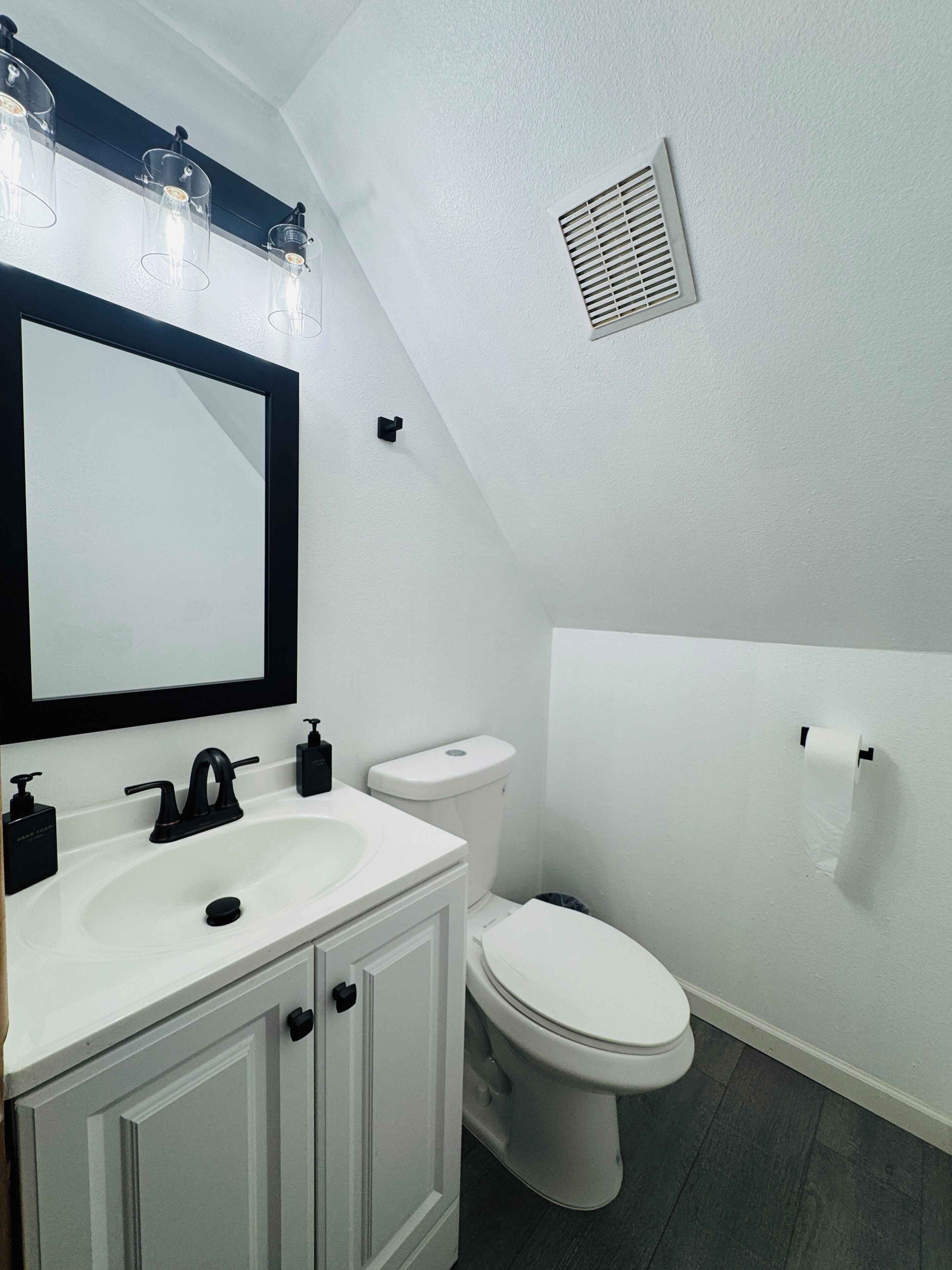 A bathroom with a white sink, black faucet, and a mirror with lights above. There is a toilet and a vent on the wall.