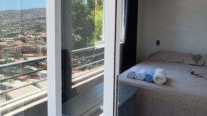 Ett rum med en säng, ett litet skrivbord och utsikt över en stadssiluett genom ett stort fönster.