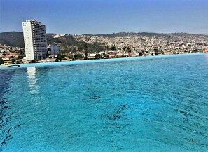 A coastal cityscape with a clear blue sea, a tall building, and a densely populated hillside in the background. - 2Exclusive, fully furnished apartment with ocean view in a World Heritage Site (Valparaiso)