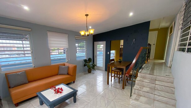 A living room with a sofa, coffee table, dining table, and chairs. There are windows with blinds, a ceiling fan, and a staircase.