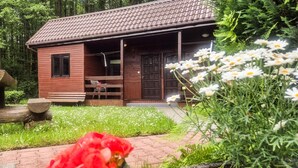 A wooden cabin with a porch, surrounded by a garden with white and red flowers, and a forested background. - 1 bedroom gorgeous home in Lazy (Lazy)