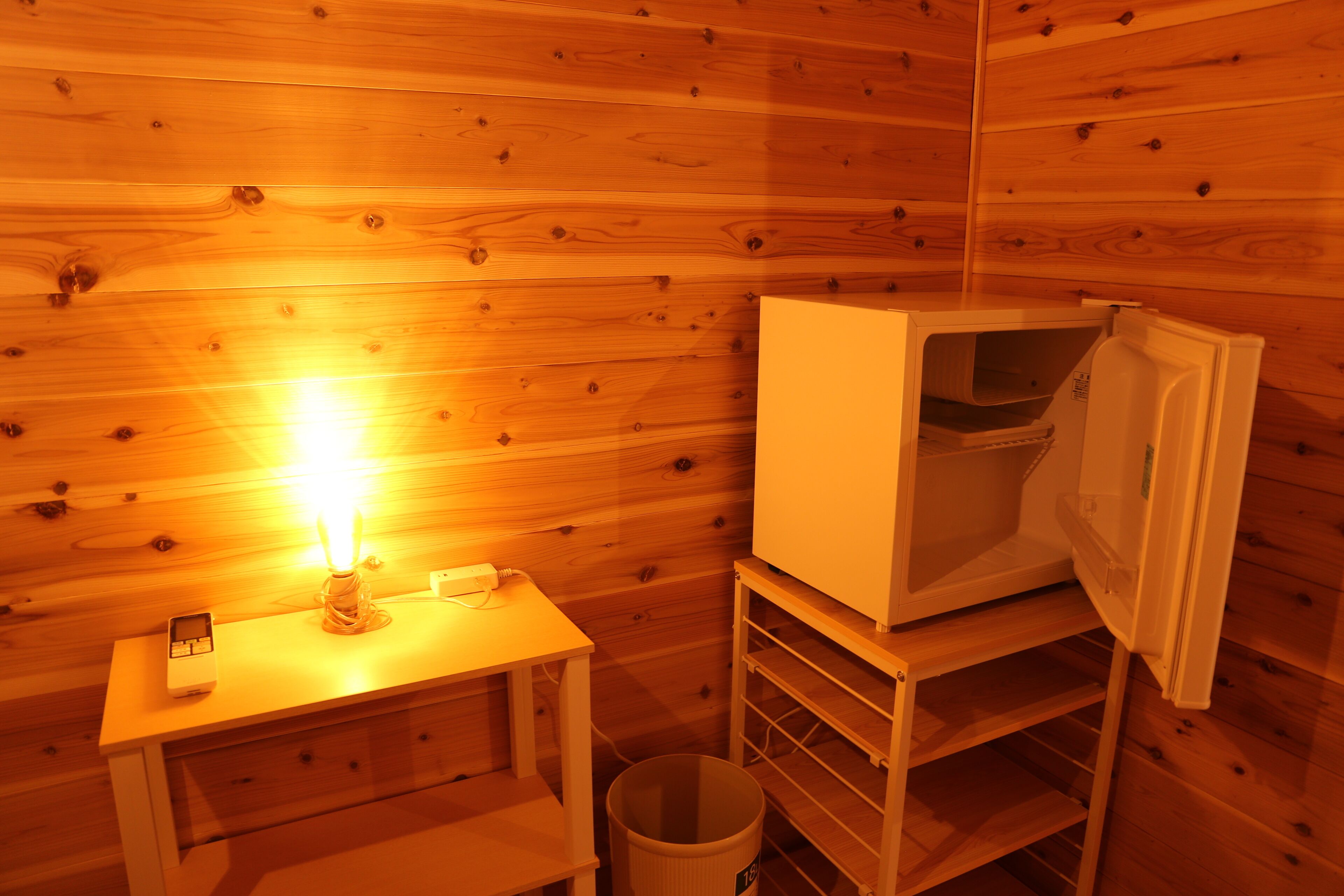 Interior de una cabaña de madera con una mesa pequeña, una vela encendida, un cesto de basura y un armario blanco.