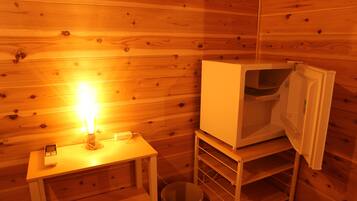 Interior de una cabaña de madera con una mesa pequeña, una vela encendida, un cesto de basura y un armario blanco.