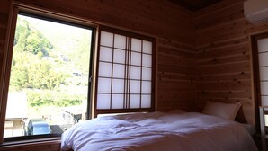 A wooden cabin room with a bed, a large window with a view of a forested area, and a small table with a chair. - Riviera Iya Valley (Miyoshi)