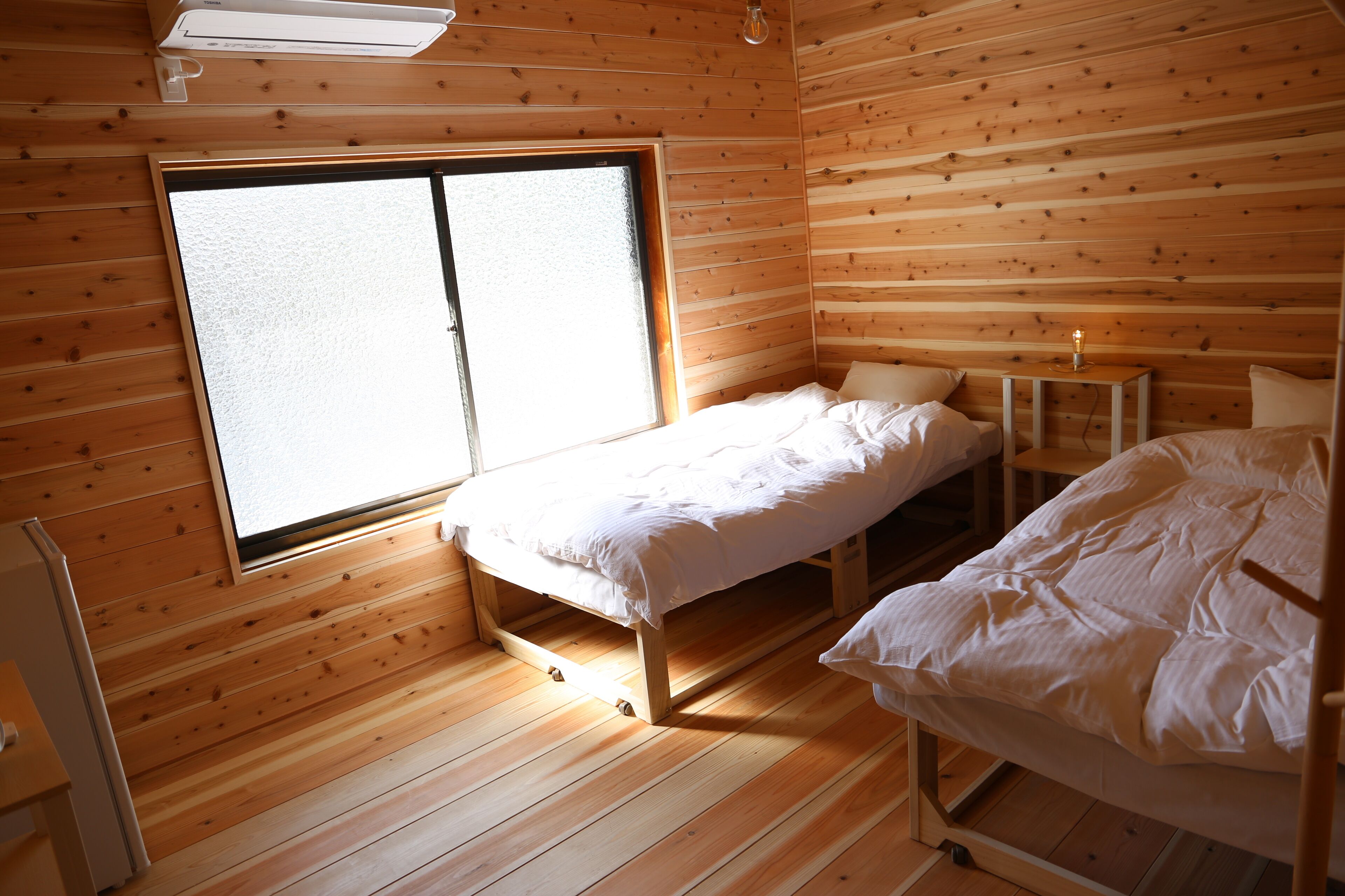 Una pequeña habitación de cabaña de madera con dos camas, una ventana y una mesita con lámpara.