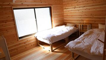 Una pequeña habitación de cabaña de madera con dos camas, una ventana y una mesita con lámpara.