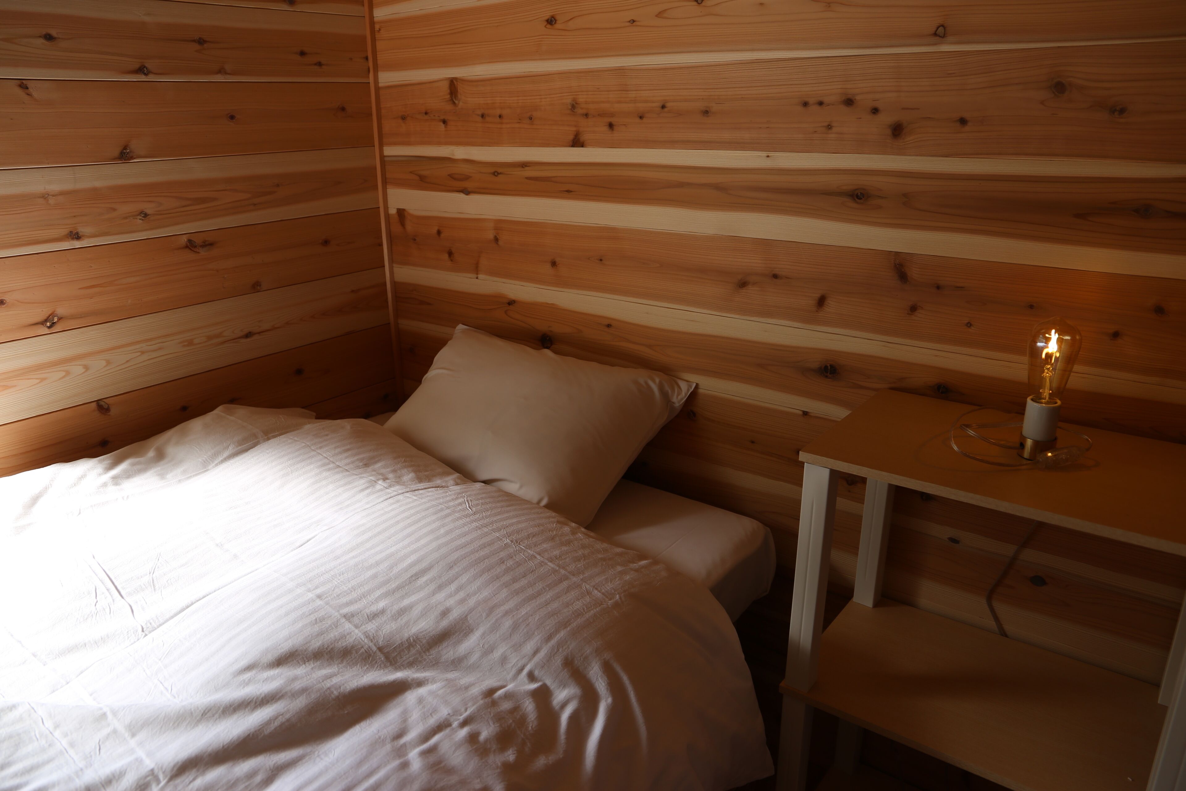 Quarto com paredes de madeira, cama com roupa de cama branca e mesa de cabeceira com uma vela.