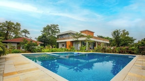 Ein Swimmingpool mit Fliesenveranda, umgeben von üppiger Bepflanzung und einem modernen Gebäude mit Balkon.
