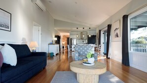 Ein modernes Wohnzimmer mit einem dunklen Sofa, einem runden Couchtisch und einem gemusterten Sessel. Im Hintergrund ist eine Küchenzeile mit Backsteinwand und Pendelleuchten zu sehen.