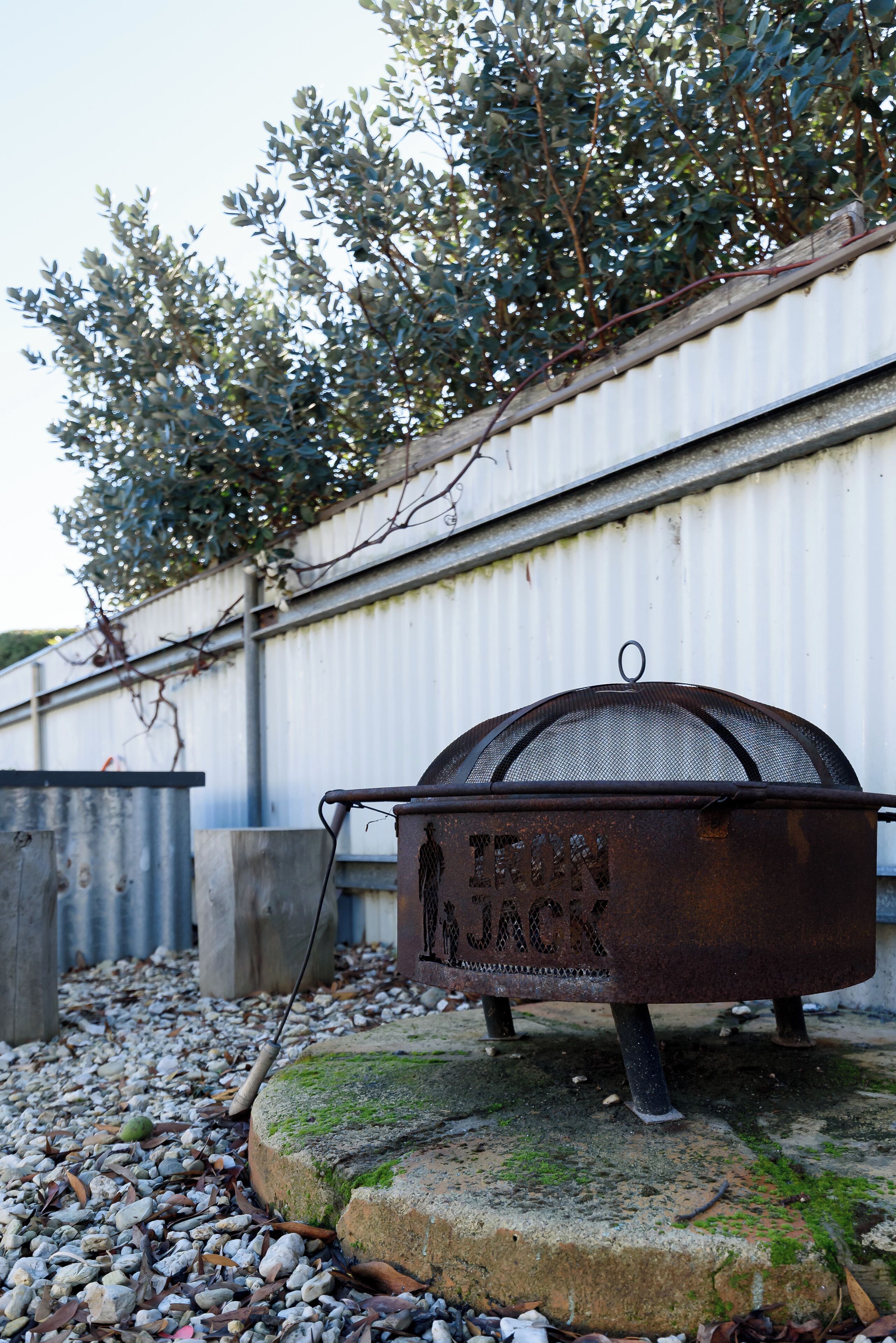 Ein Metallgrill mit der Aufschrift "IRON JACK" auf einem Steinsockel, vor einer wellenförmigen Metallwand und einem Baum mit grünen Blättern.