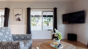 Ein Wohnzimmer mit einem gemusterten Sofa, einem runden Couchtisch mit Blumenvase, einem Flachbildfernseher und einem Fenster mit Vorhängen.