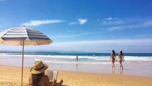 A beach scene with a striped umbrella, a person reading a book, and two individuals walking on the sand. - Culburra Beach B&B Jervis Bay Area Private Room Shared bath with Jacuzzi Pool AC (Culburra Beach)