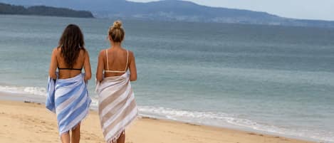 Deux personnes se promènent sur une plage de sable, avec vue sur l’océan et les collines au loin.