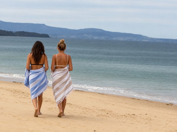 Two individuals walking on a sandy beach with a view of the ocean and distant hills.