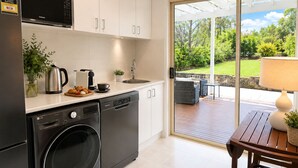 A modern kitchen with a dishwasher, microwave, and washing machine. There is a dining area with a table and chairs, and a view of a garden through a glass door. - Culburra Beach B&B Jervis Bay Area Private Room Shared bath with Jacuzzi Pool AC (Culburra Beach)