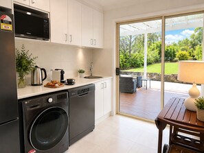 Eine moderne Küche mit Geschirrspüler, Mikrowelle und Waschmaschine. Ein Essbereich mit Tisch und Stühlen bietet Blick auf einen Garten durch eine Glastür.