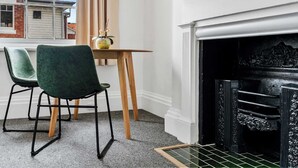 Een kamer met een open haard, twee stoelen, een kleine tafel met een vaas en een raam met uitzicht op een gebouw.