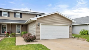 A two-story house with a white garage door, beige siding, and a front yard with grass and shrubs.
