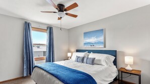 A bedroom with a bed, a ceiling fan, a nightstand with a lamp, a window with curtains, and a painting on the wall.