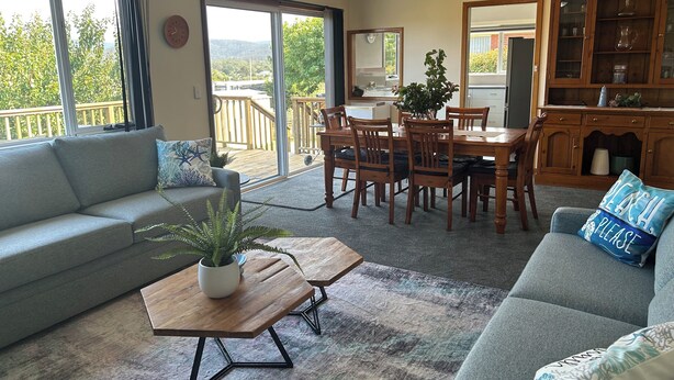 Un vaste salon doté d’un canapé gris, d’une table basse en bois et d’un coin repas avec une table et des chaises en bois. Une grande porte-fenêtre coulissante donne accès à un balcon avec une terrasse en bois.