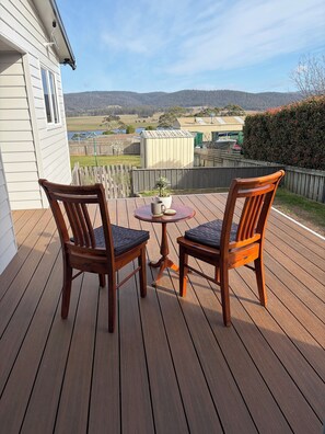 A wooden deck with two chairs and a small table, overlooking a rural landscape with hills and a fenced area. - Cosy Coastal Cottage close to Maria Island Ferry - recently renovated (Triabunna)