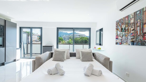 A modern hotel room with a large bed, two sofas, a glass door leading to a balcony, and a cityscape view. - Central 1-Br with Balcony – Rue d’Antibes, Cannes (Cannes)