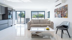 A modern living room with a sofa, a coffee table, and a chair. There is a kitchen area with cabinets and an oven. A large window provides a view of the outdoors. - Central 1-Br with Balcony – Rue d’Antibes, Cannes (Cannes)
