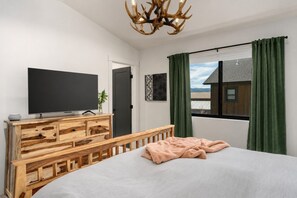 Une chambre à coucher avec un lit, un téléviseur posé sur une commode en bois, une fenêtre avec des rideaux et une vue sur un bâtiment à l’extérieur.