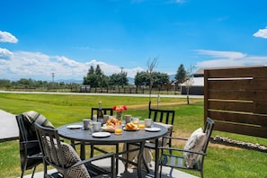 A round outdoor table with chairs, a bottle, a vase with flowers, and breakfast items.