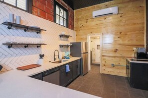 A modern kitchen with a herringbone tile backsplash, wooden wall paneling, a stainless steel refrigerator, and a built-in dishwasher. - Restored Historic Loft - 3 Mins to the Beach (Galveston)