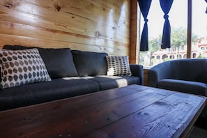 A wooden wall, a sofa with patterned pillows, a wooden coffee table, and a view of buildings through a window. - Restored Historic Loft - 3 Mins to the Beach (Galveston)